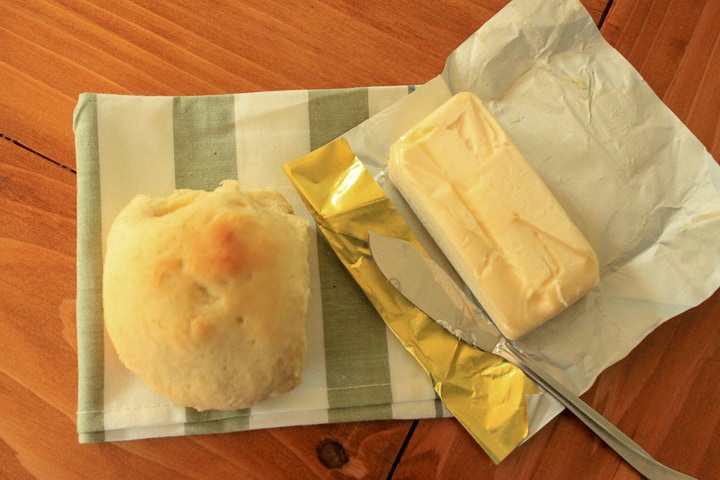 single vegan dinner roll on a striped napkin with a stick of vegan butter and butter knife all sitting on a wood background. 