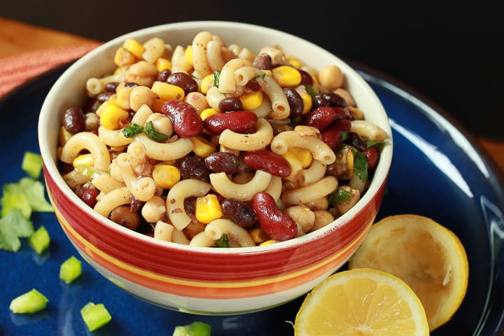 Mexican Pasta Salad in a colorful bowl on a dark blue plate with a sliced lemon and peppers. 