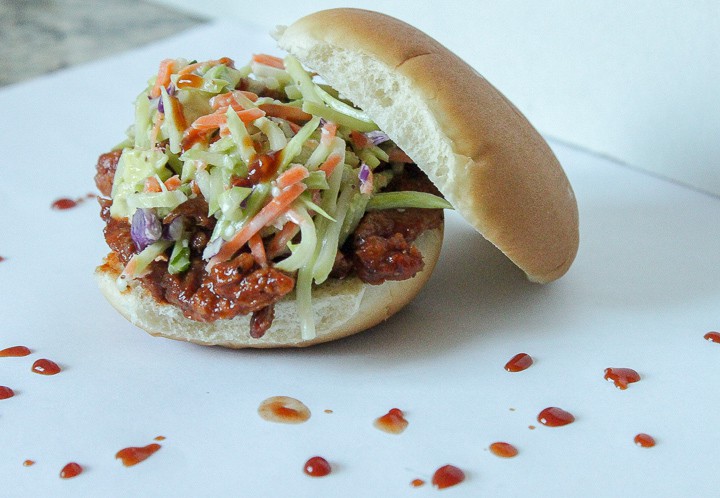 vegan pulled pork sandwich with brocolli slaw on a white background with half the bun off and drops of bbq sauce around it. 