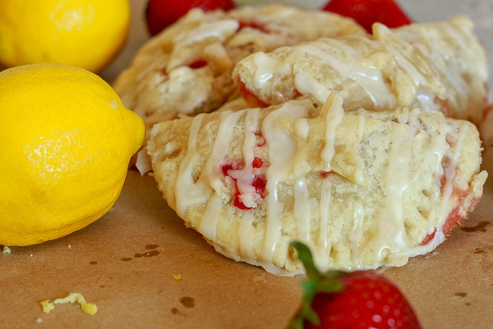 A pile of vegan Hand Pies cooked with drizzle on them on a brown background with a lemon and strawberry in the photo. 