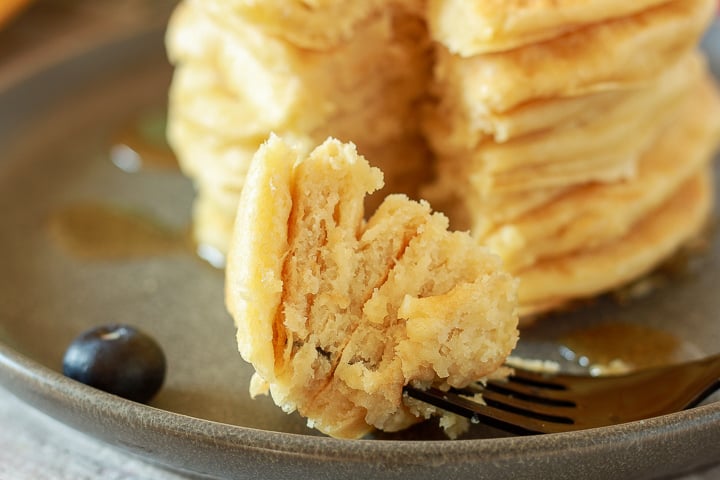 fluffy vegan pancakes bite on a fork with 7 stacks of a pancake on a black plate. 