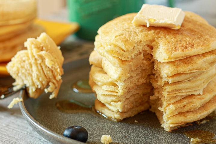 a stack of vegan pancakes with vegan butter on top and syrup and bite taken out with a fork on a black plate. 