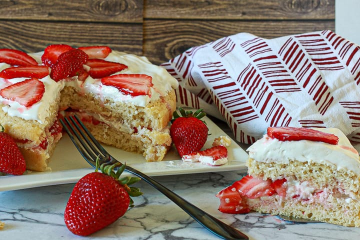 vegan strawberry shortcake with the whole cake with a slice out of it in the background and a slice in the front with a fork on a marble background.