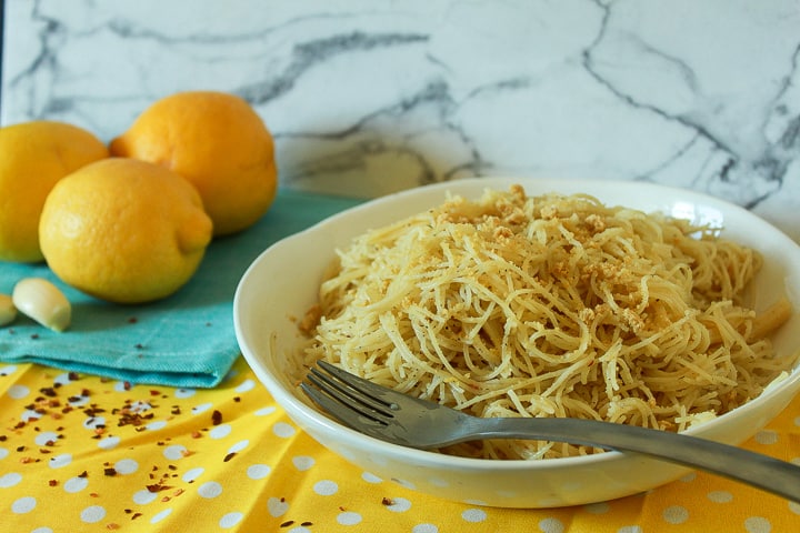 vegan garlic lemon pasta in a white bowl with a fork on a polka dot yellow background with lemons and garlic pictured.