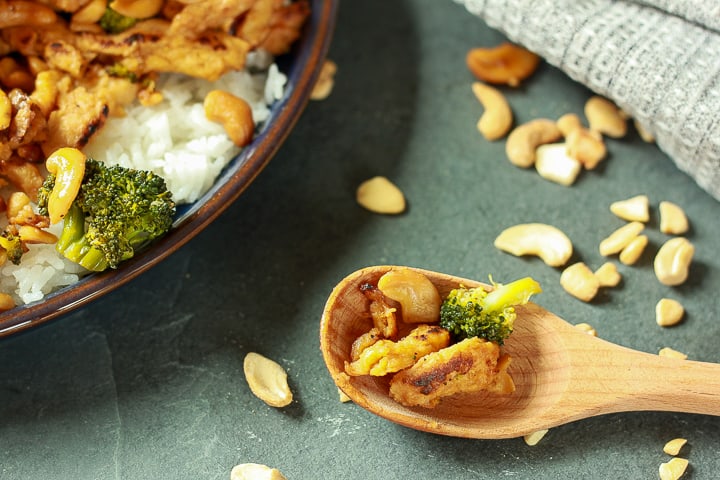 vegan cashew chicken on a wooden spoon with a portion of the main dish showing and cashews sprinkled around it. 