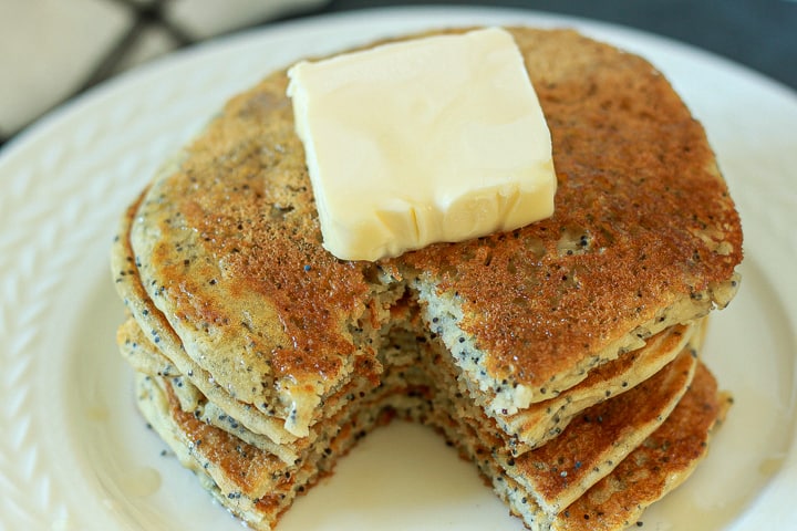 almond poppyseed pancake stack with a stack of butter on top and syrup on a white plate. 