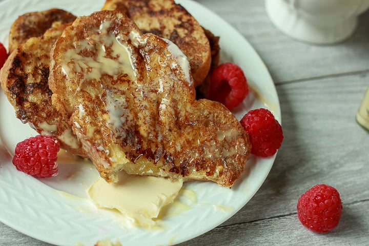 french toast plated on a white plate with maple butter, vegan butter, and raspberries on a white wood background. 