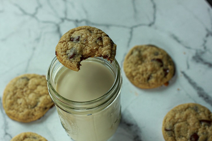 Vegan Chocolate Chip Cookies on a marble background surrounding a glass of milk with a bite taken out of a cookie on the rim of the glass of milk. 