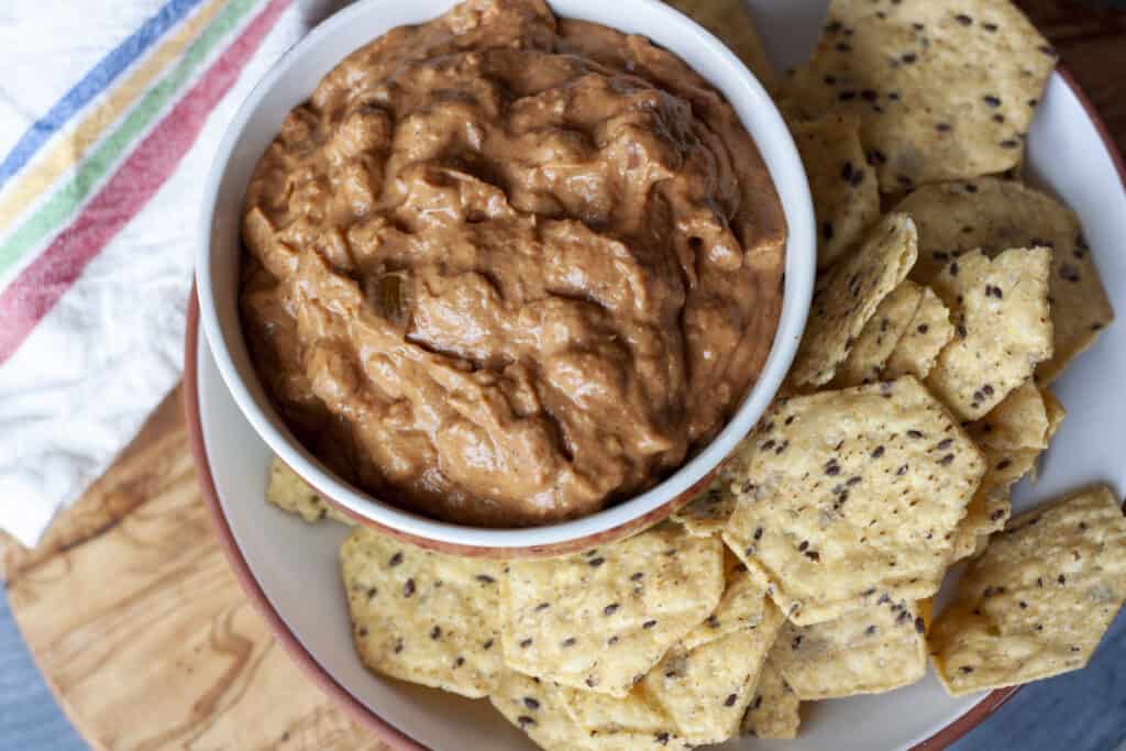 Vegan bean dip in a small bowl with tortilla chips around it on a plate then on a wood background. 