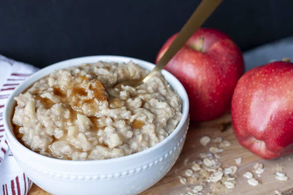 vegan apple cinnamon oatmeal filled to the top of a small white bowl with some brown sugar melted on top with a gold spoon and two apples in the background. 