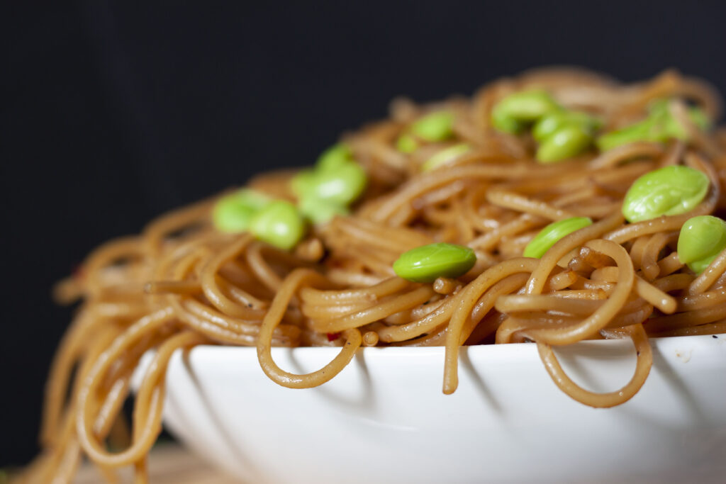 hibachi noodles in a white bowl with a black background and edamame scattered throughout. 