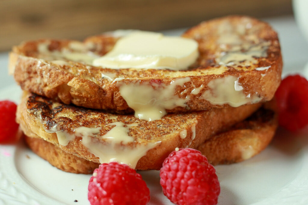 3 stacks of vegan french toast with maple butter drizzled on and raspberries scatterd on a wite plate. 