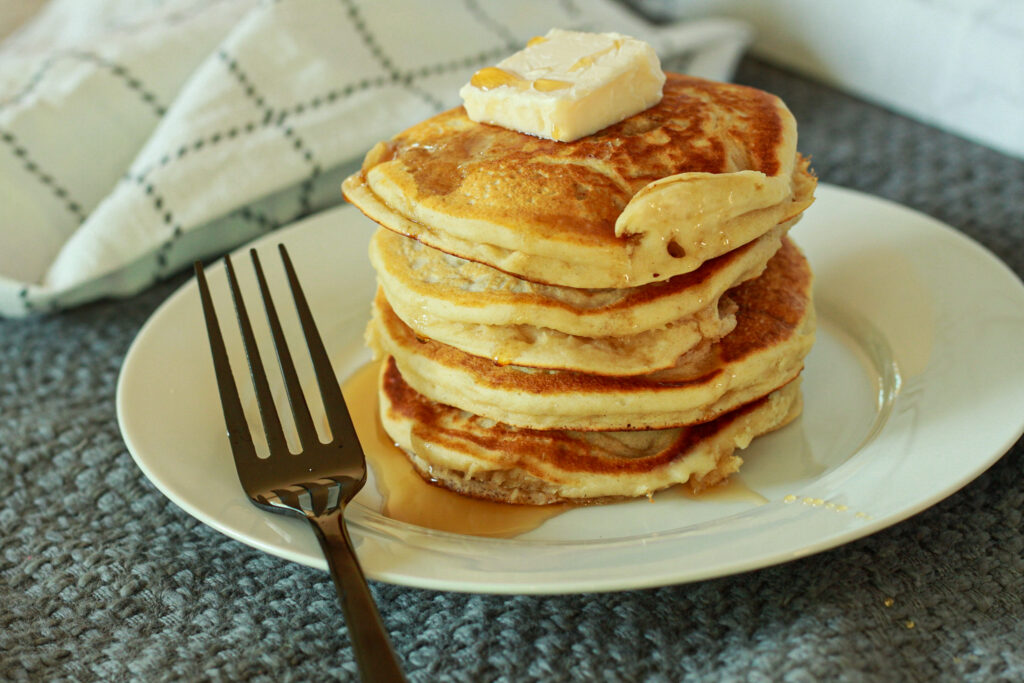 vegan buttermilk pancakes 4 pancakes high with a slab of vegan butter on top with some syrup on a white small plate. 