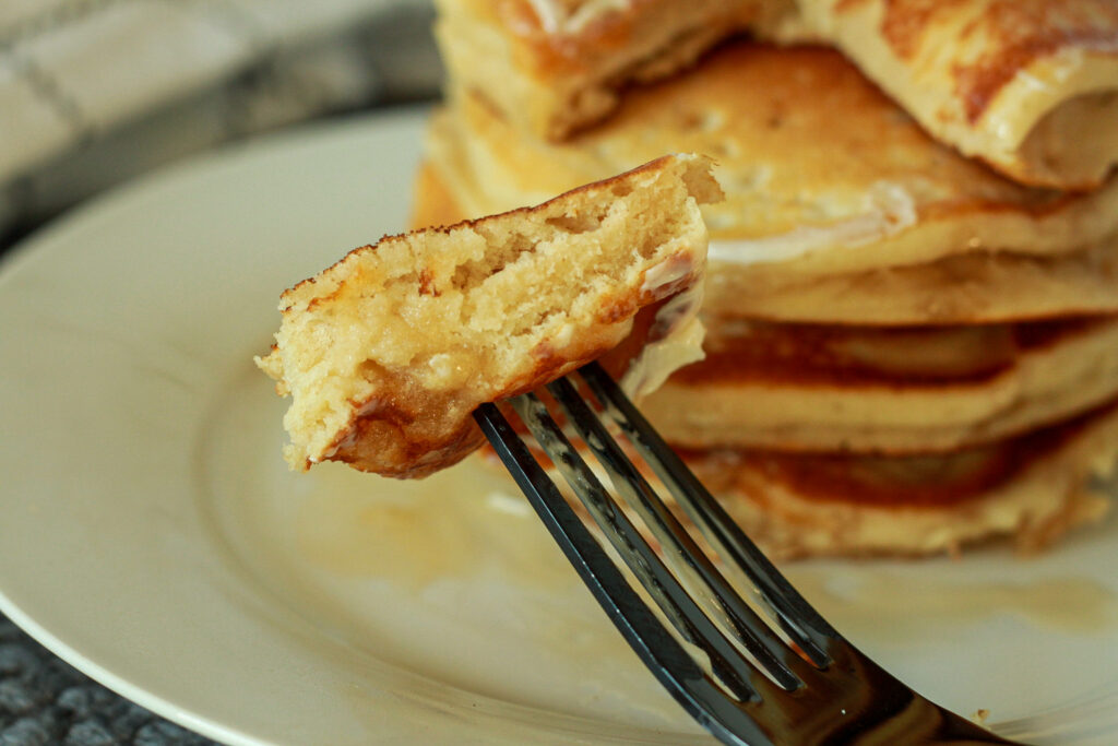 fluffy vegan pancake bite on a fork with a stack of pancakes on the back on a white plate. 