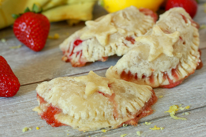 3 vegan fruit hand pies with star shaped pie crust on the top on a white wood background with strawberries in the background. 