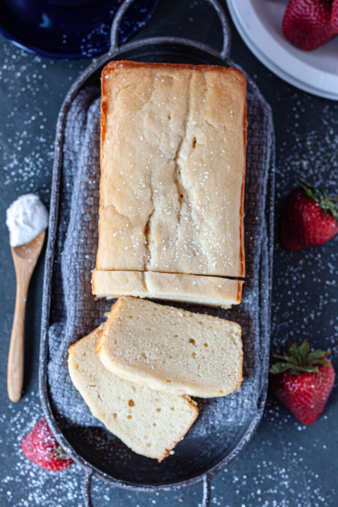 pound cake loaf with a few slices on a grey towel and strawberries on a grey background with a spoon of whipped cream to the side. 