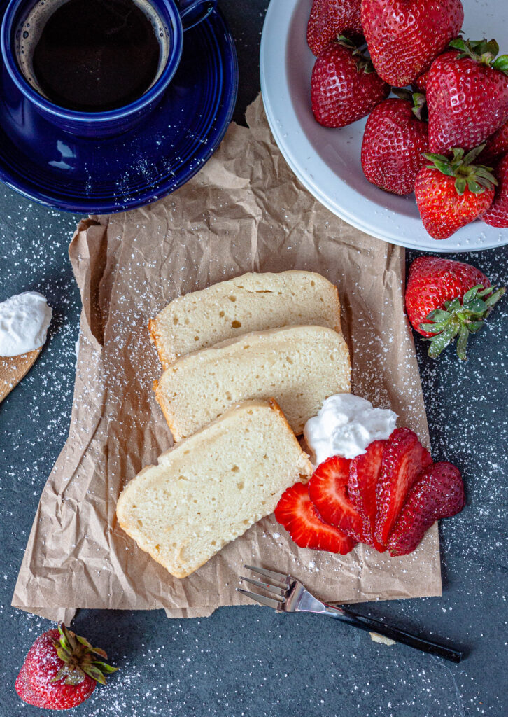 vegan pound cake slices and strawberries and whip cream around it on a brown paper bag with a grey background. 