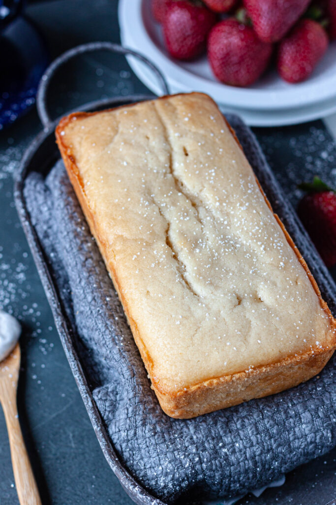 vegan pound cake loaf with a sprinkle of powdered sugar on a grey dish towel in a grey dish on a grey bakcground. 