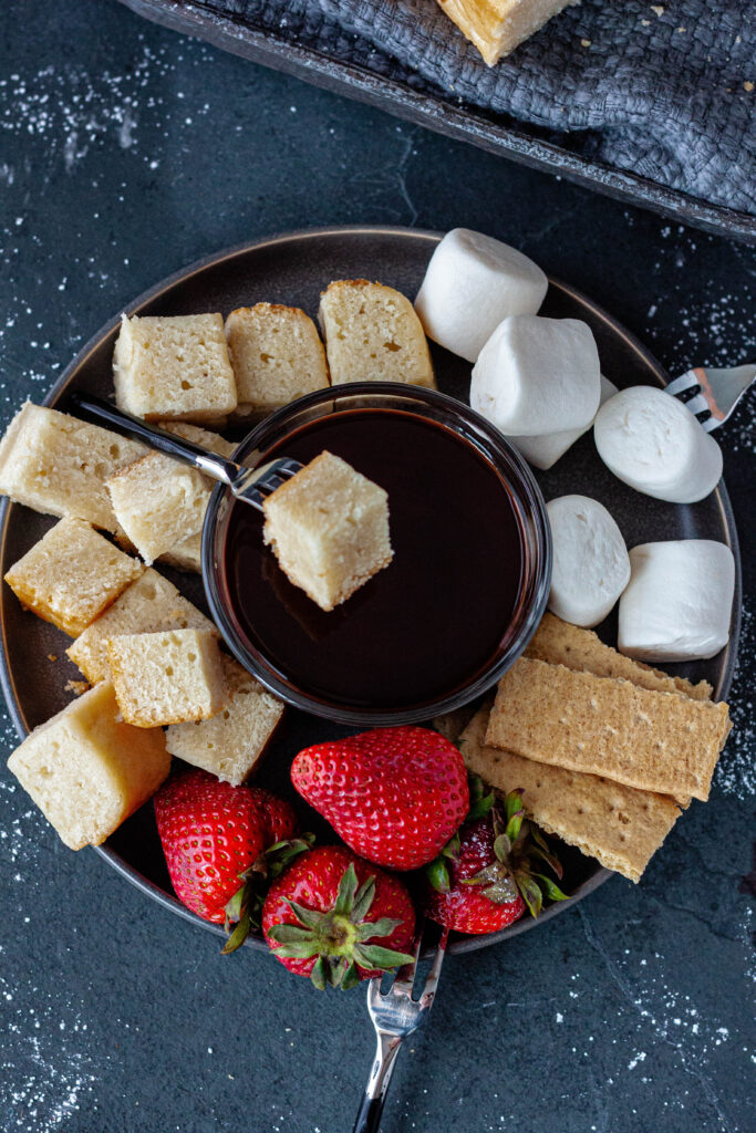 vegan pound cake fondue with a small bowl of chocolate sauce in the middle with pound cake squares, marshmallows, graham crackers, and strawberries. 