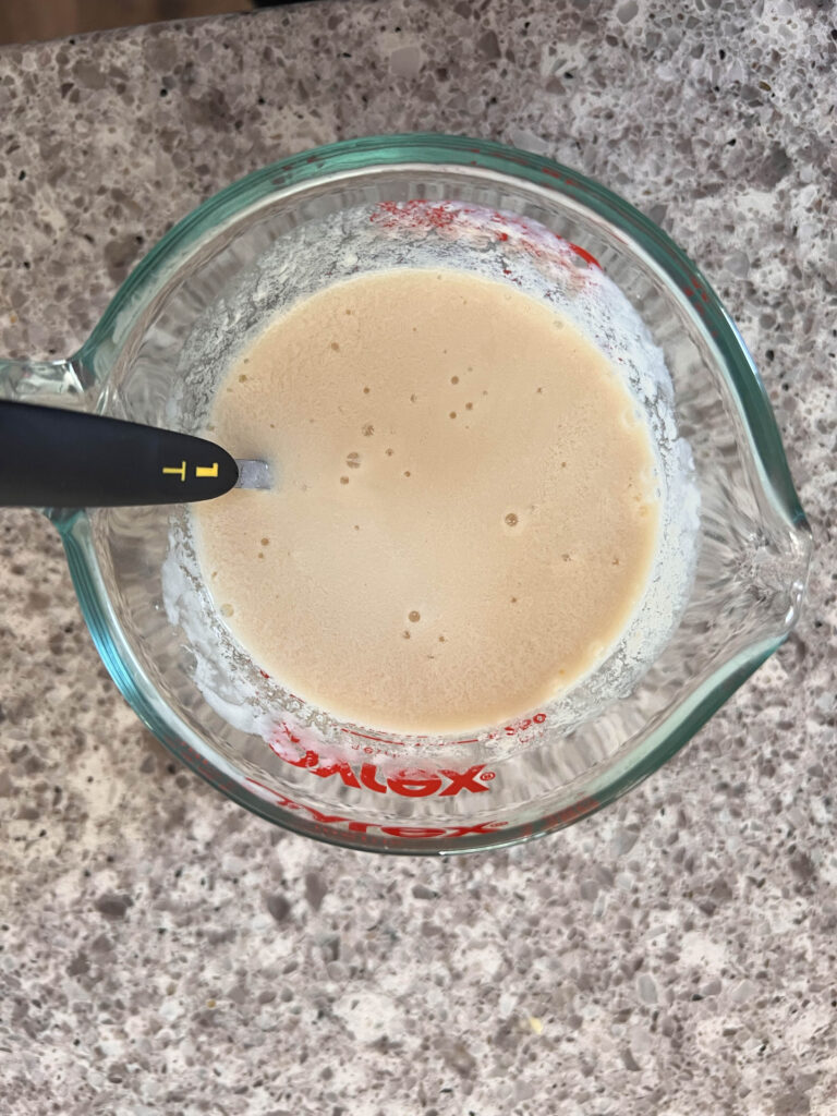 A 1 cup pyrex glass mixing cup with non dairy milk with the vinegar stirred in and 1 teaspoon measuring spoon in the cup. 