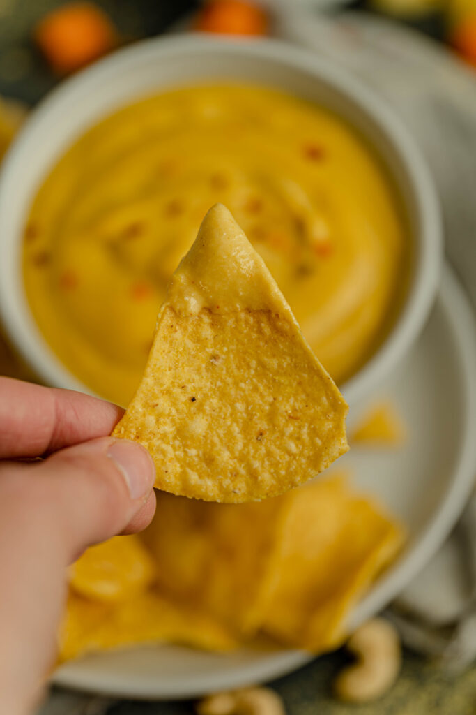 vegan nacho cheese dip in the background with a chip front and center with nacho cheese sauce on it. 