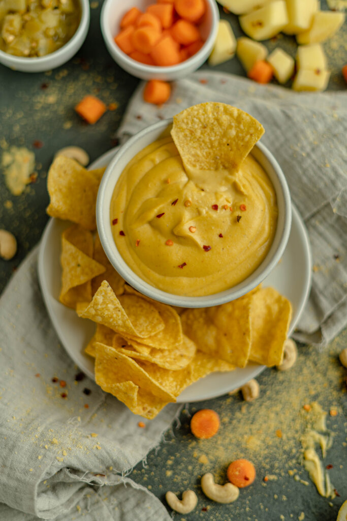 vegan nacho cheese in a bowl with chips on plate surrounding it and one chip dipped in the sauce. 