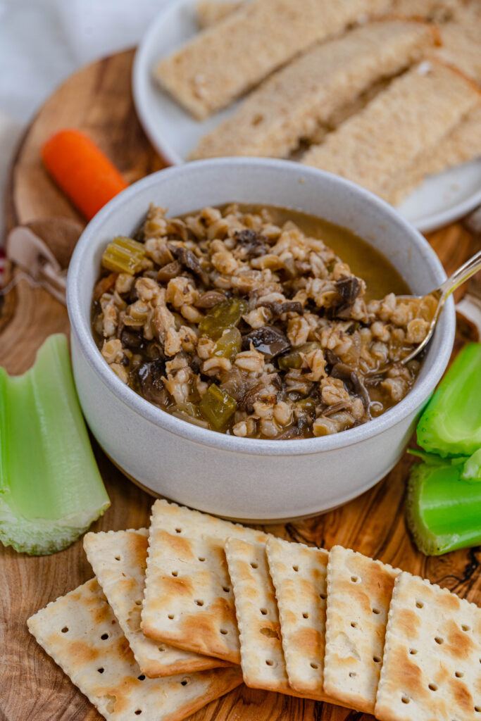 bowl of vegan barley soup on a wood background with saltine crackers and celery surrounding it. 
