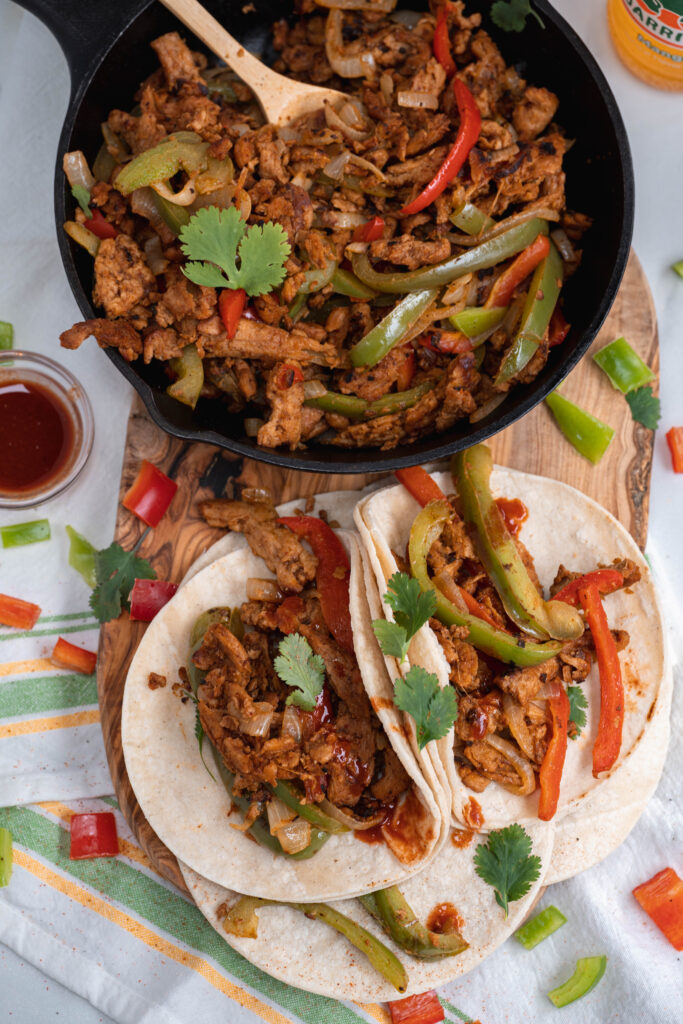 vegan cooked fajitas plated on a wood background with a skillet behind it full of the fajita mix. 