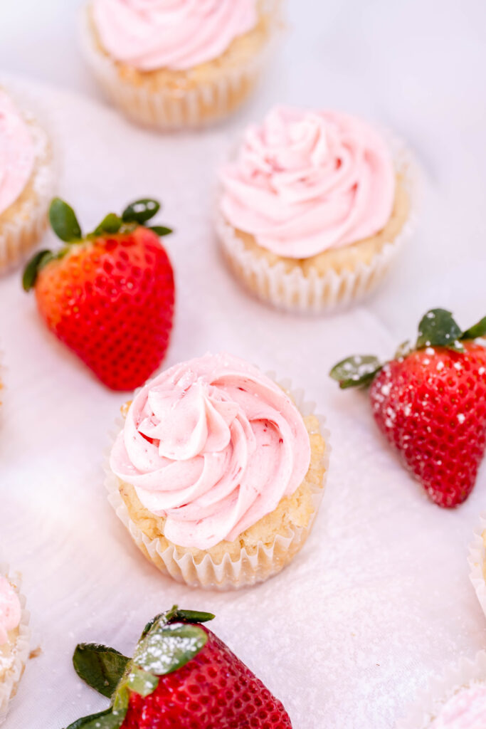 vegan strawberry shortcake cupcakes frosted on a pink sheet with strawberries to the side. 