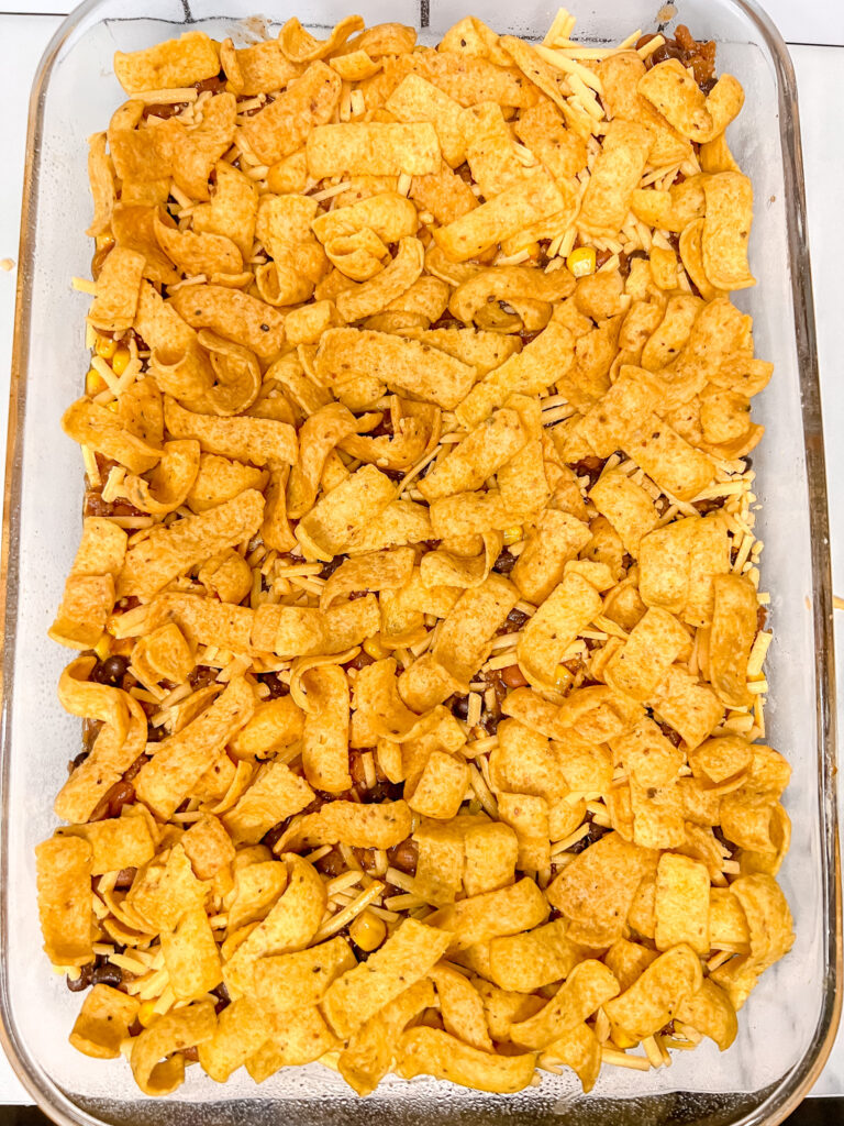 vegan frito pie covered in fritos before baking in a class pan on a white background. 