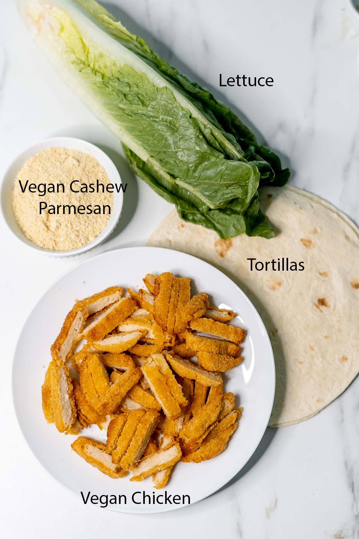 Vegan Chicken, Lettuce, Tortillas, and vegan parmesan cheese in a bowl or plates on a marble background with black lettering. 