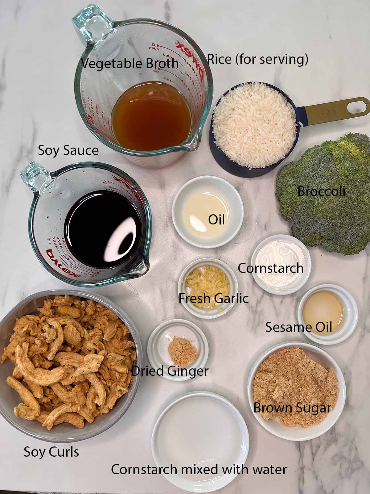 Ingredients for the vegan beef and broccoli all in small bowls with black labels on what they are on a marble background. 