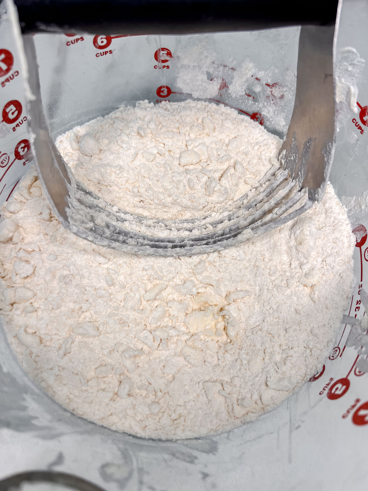 Flour mixture in a glass bowl with little pieces of vegan butter and a pasty cutter in the flour mixture. 