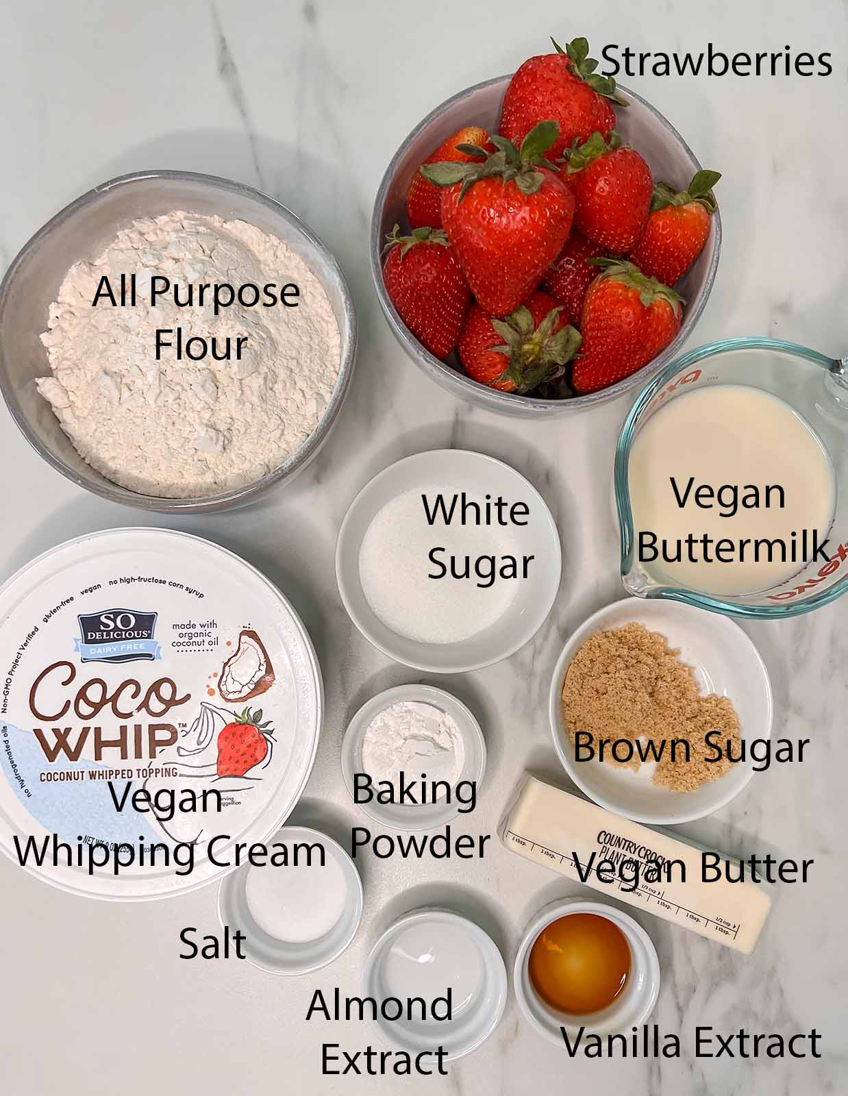 Vegan strawberry shortcake ingredients all in small bowls or containers with black labels on what they are on a marble background. 