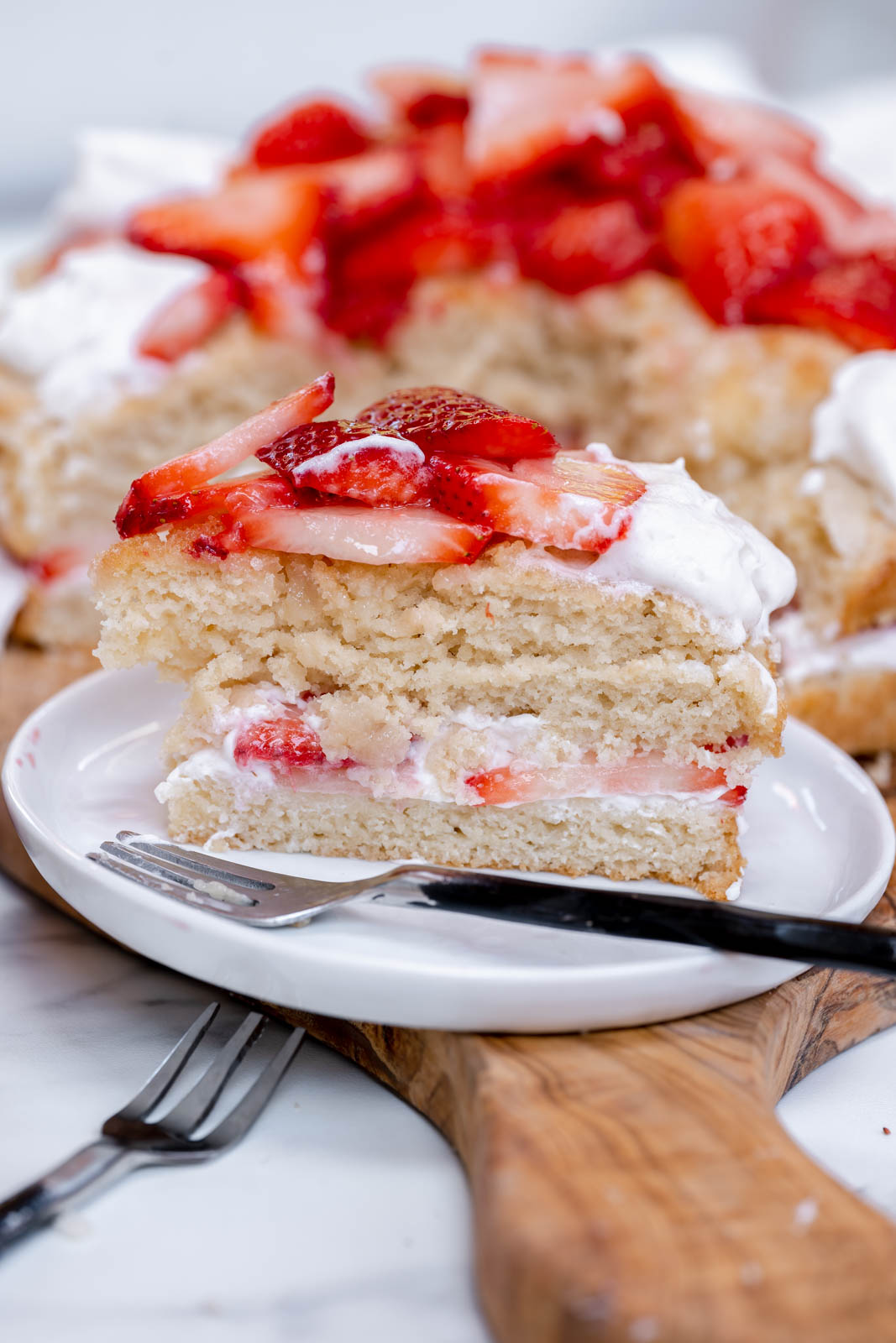 A single slice of vegan strawberry shortcake on a round white plate with a small fork on a wood piece. 