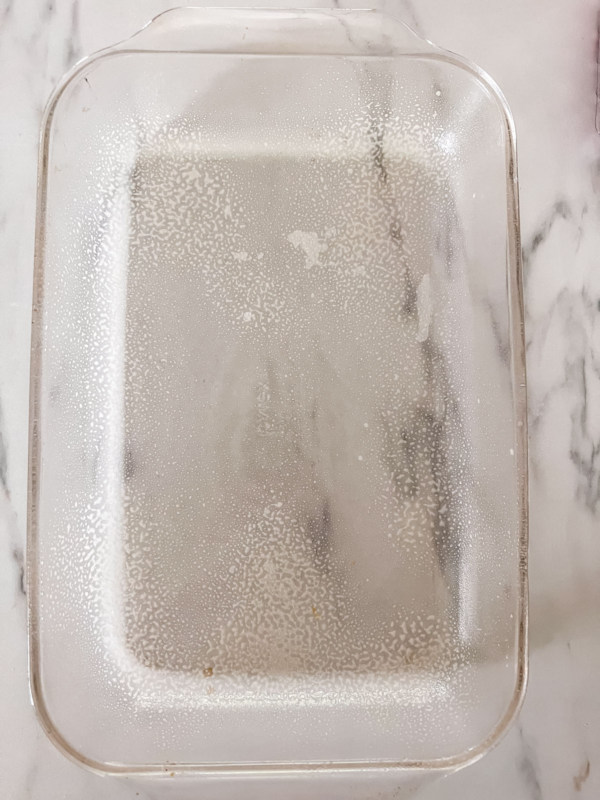A 9x13 glass pan with cooking spray in it on a marble background.n