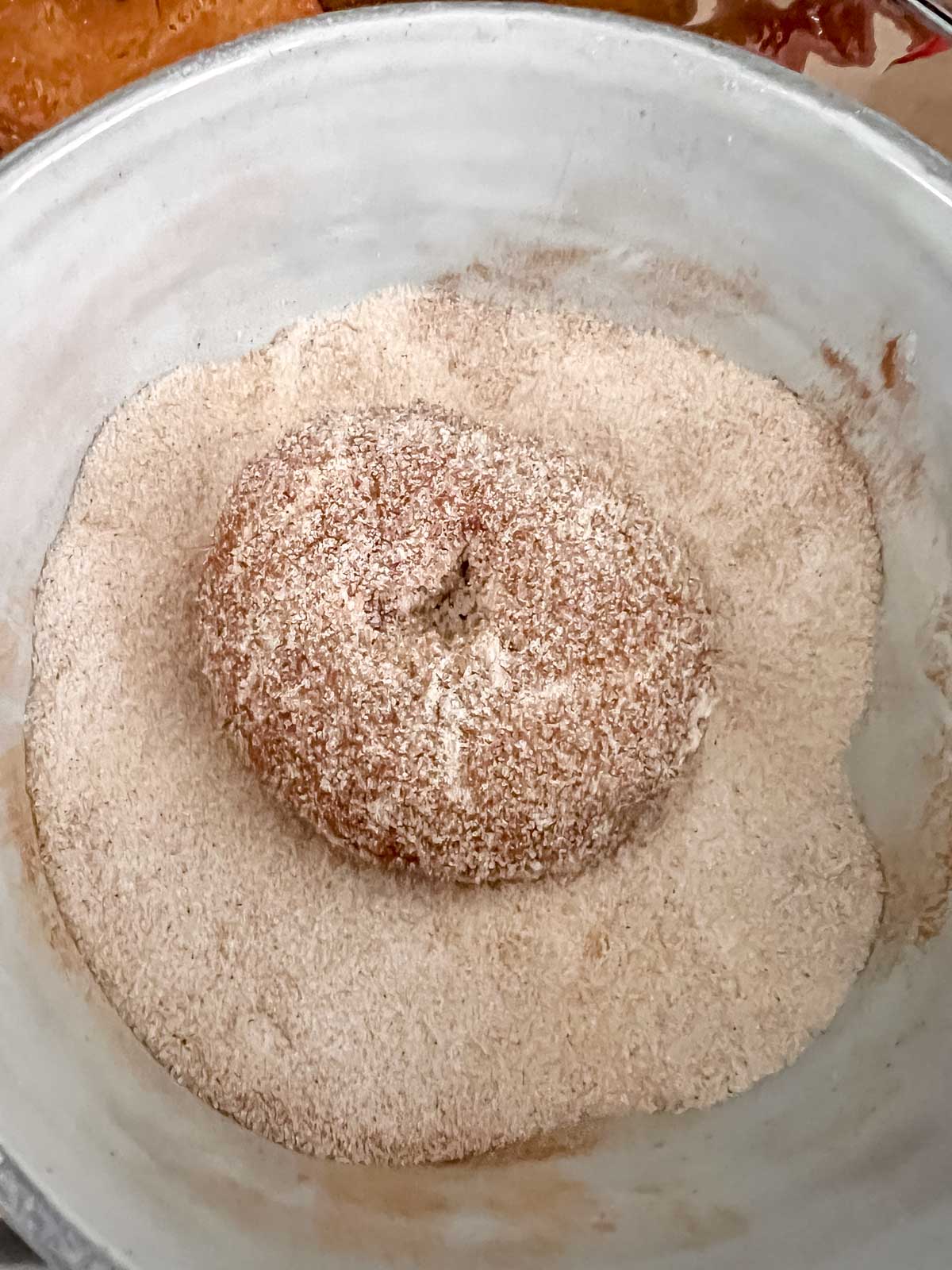 One vegan donut being dipped in the bowl of Cinnamon and sugar. 