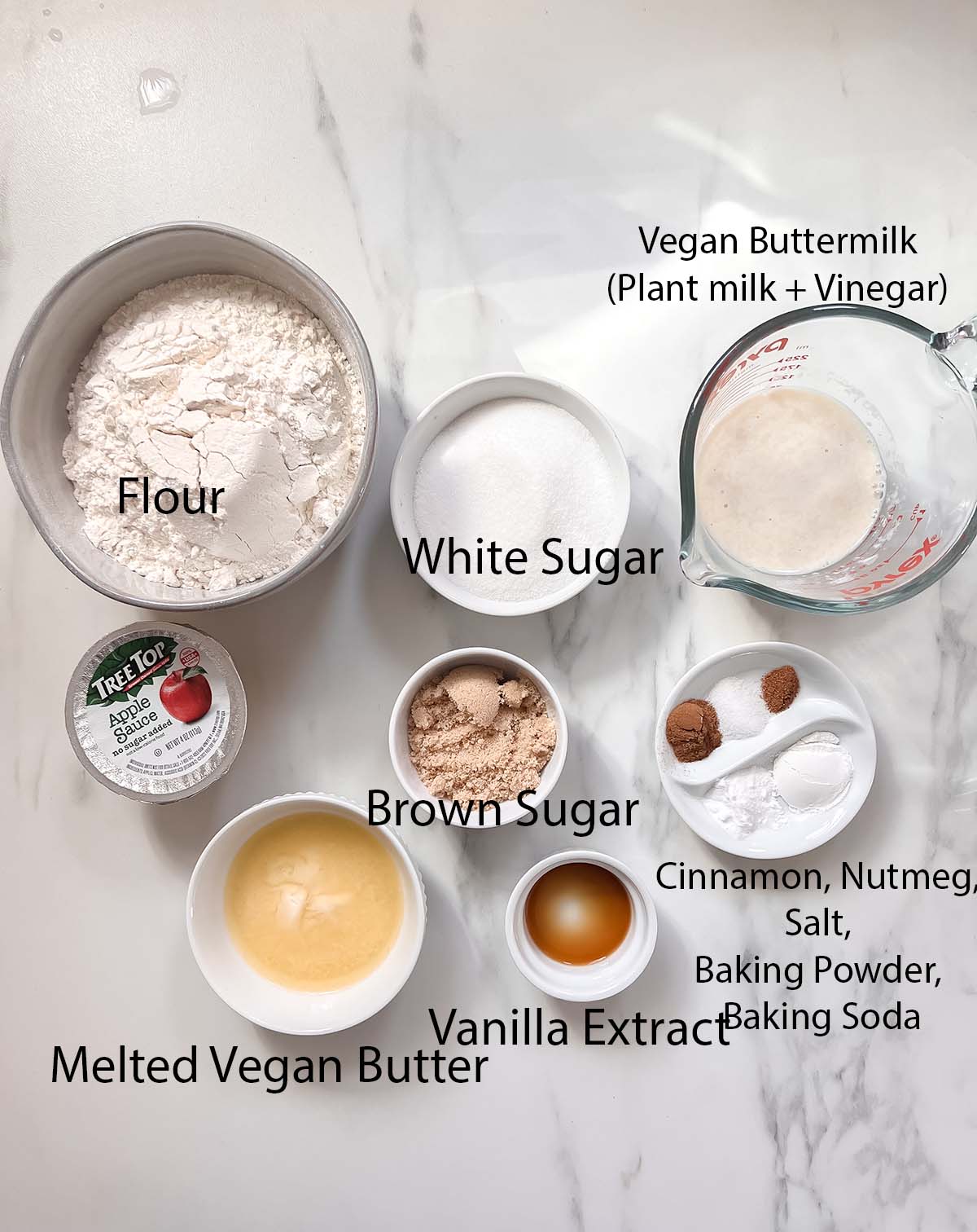 Vegan apple cider donuts ingredients in small bowls with black labels on a marble background. 