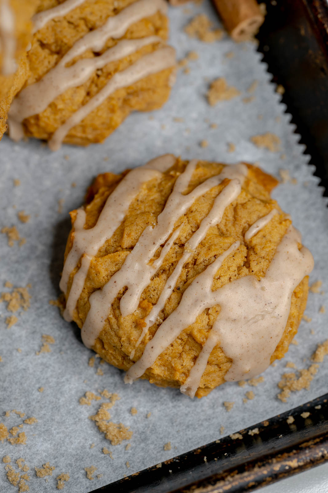 Single vegan pumpkin cookie with cinnamon drizzle on a white paper with crumbs around it. 