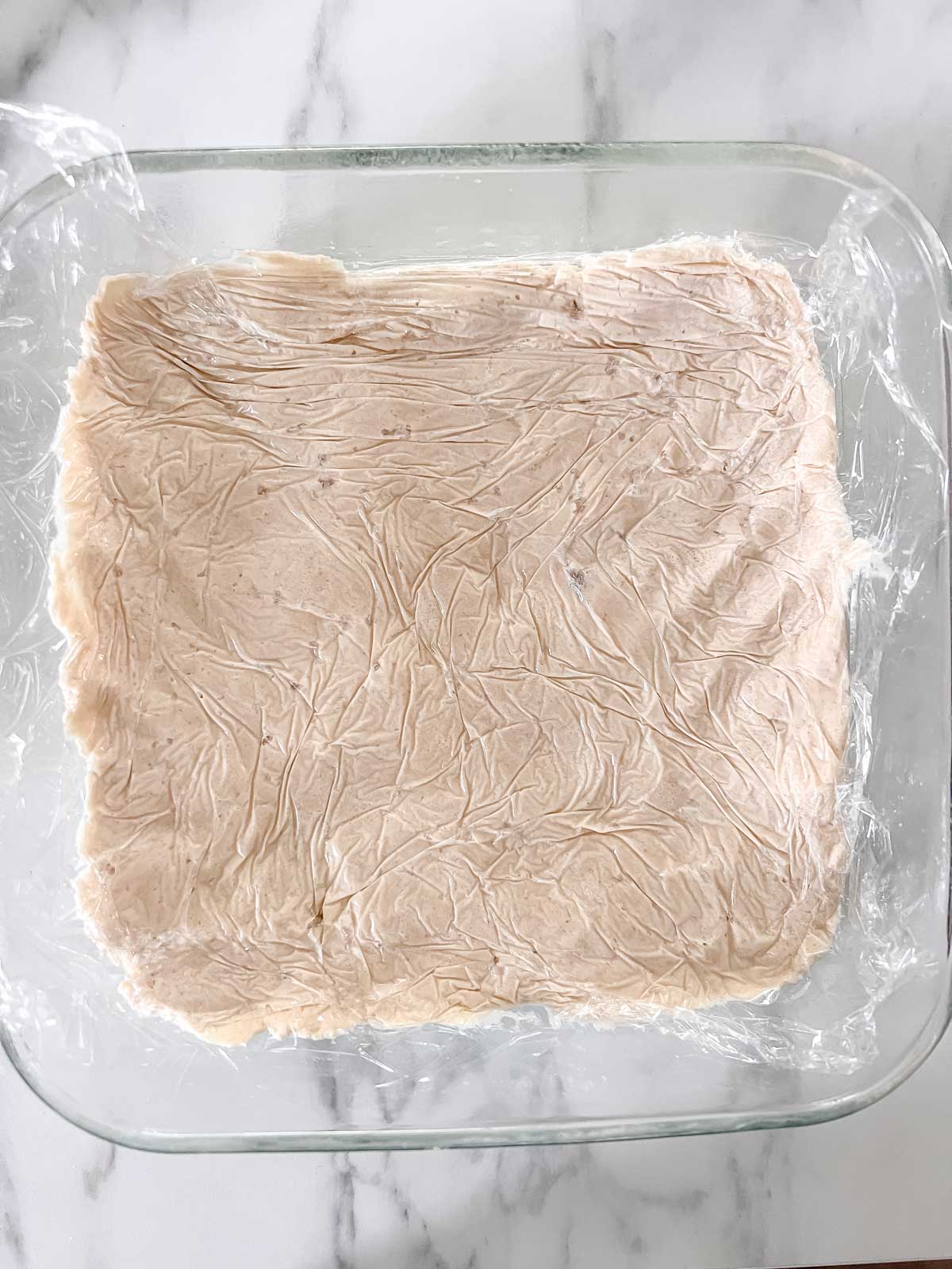 An 8x8 glass pan with half the dough pressed in it with plastic wrap over it. 