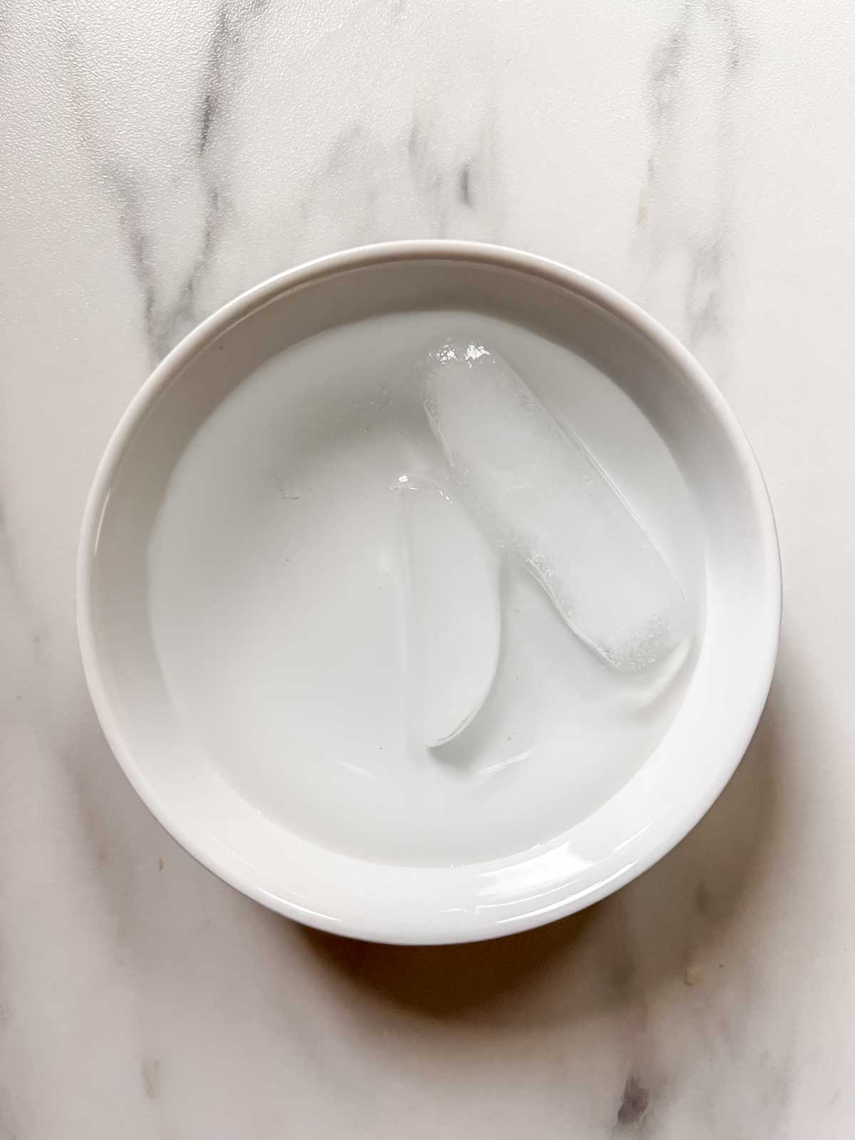 A small white bowl with water and two melting ice cubes in it on a marble background. 