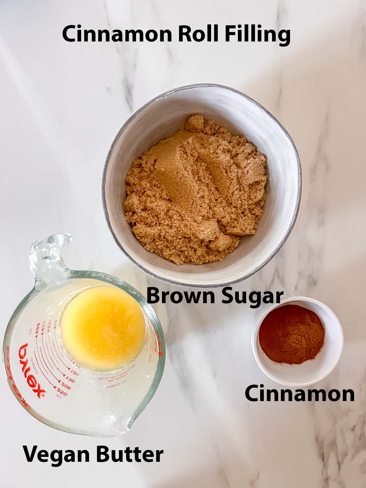 Vegan cinnamon roll filling ingredients in small bowls with black labels on a marble background. 
