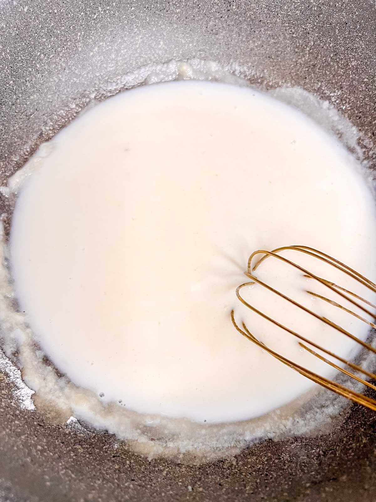 A white milky mixture being stirred with a whisk in a grey pot. 