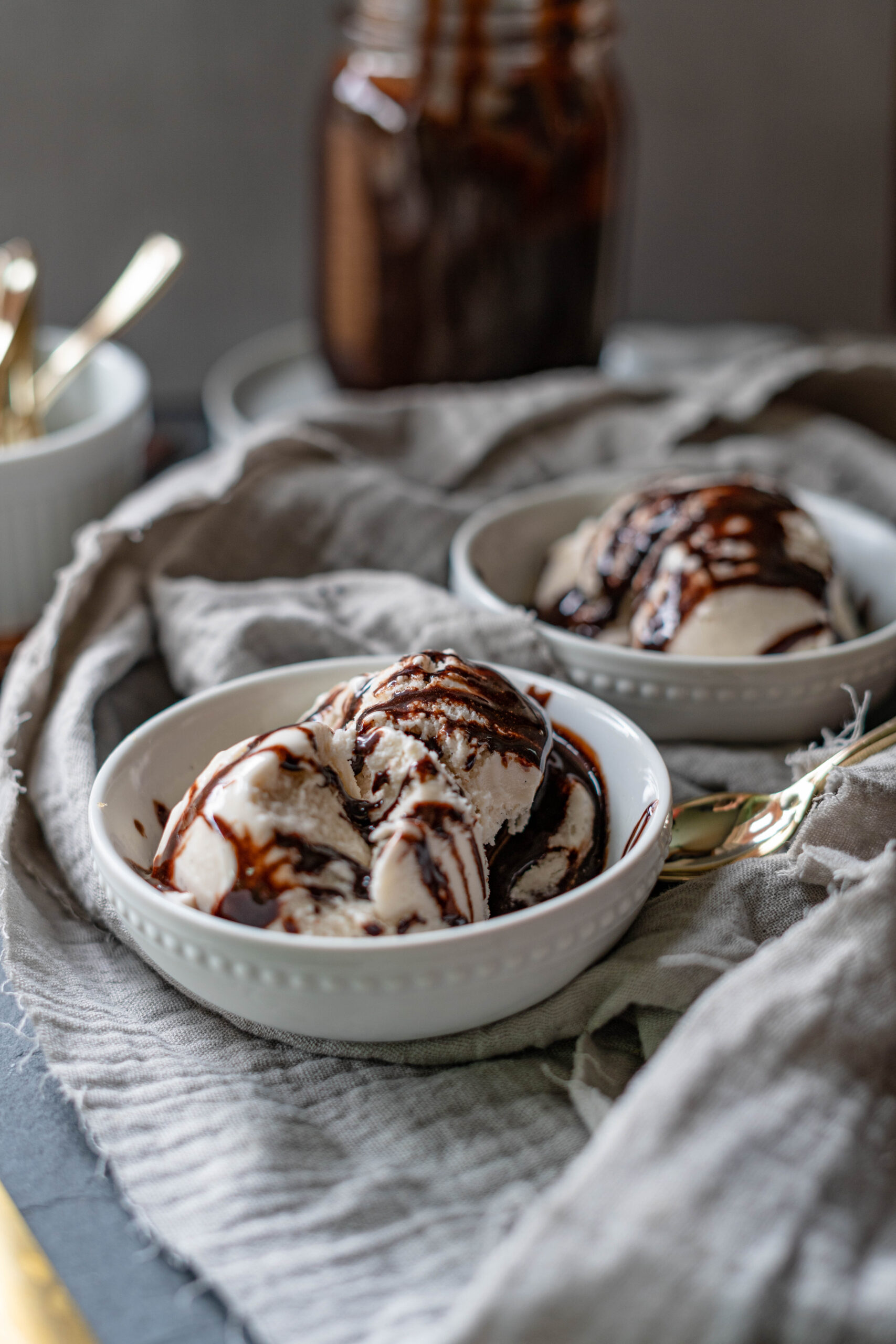 Two bowls of ice cream with vegan chocolate sauce drizzled on them on a grey fabric. 