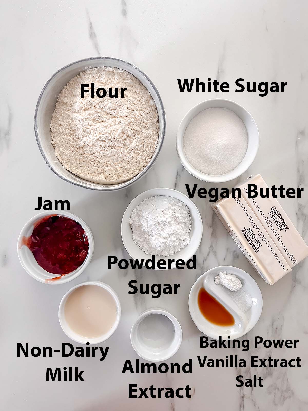 Ingredients for vegan thumbprint cookies in small bowls with black labels on a marble background. 