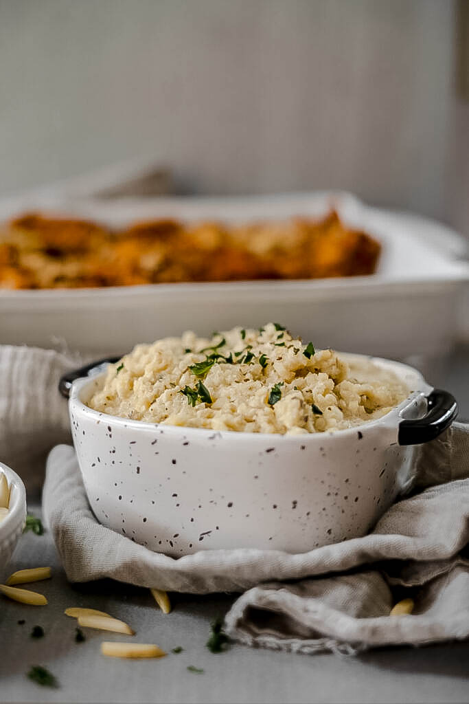 Side angle of the vegan ricotta cheese in a small black and white container with a casserole blurred out in the background. 